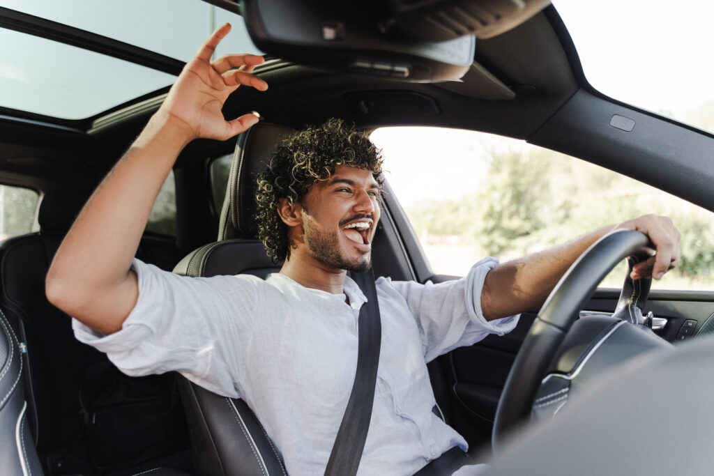 Young Indian man happily drives, singing to music with the sunroof open, enjoying a carefree road trip daydreaming of new adventures